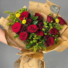 Bouquet of red and burgundy roses with greenery, wrapped in brown paper, held by a hand against a gray background.