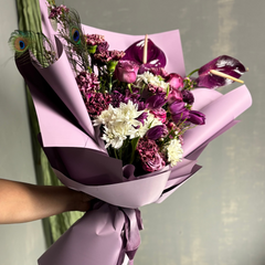 Bouquet of flowers wrapped in purple paper held by a person, with a neutral background.
