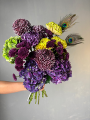 Bouquet of purple and yellow flowers with peacock feathers held by a person against a gray background