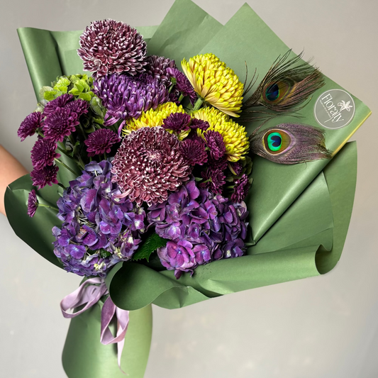 Bouquet of purple and yellow flowers with green wrap and peacock feathers on a light gray background