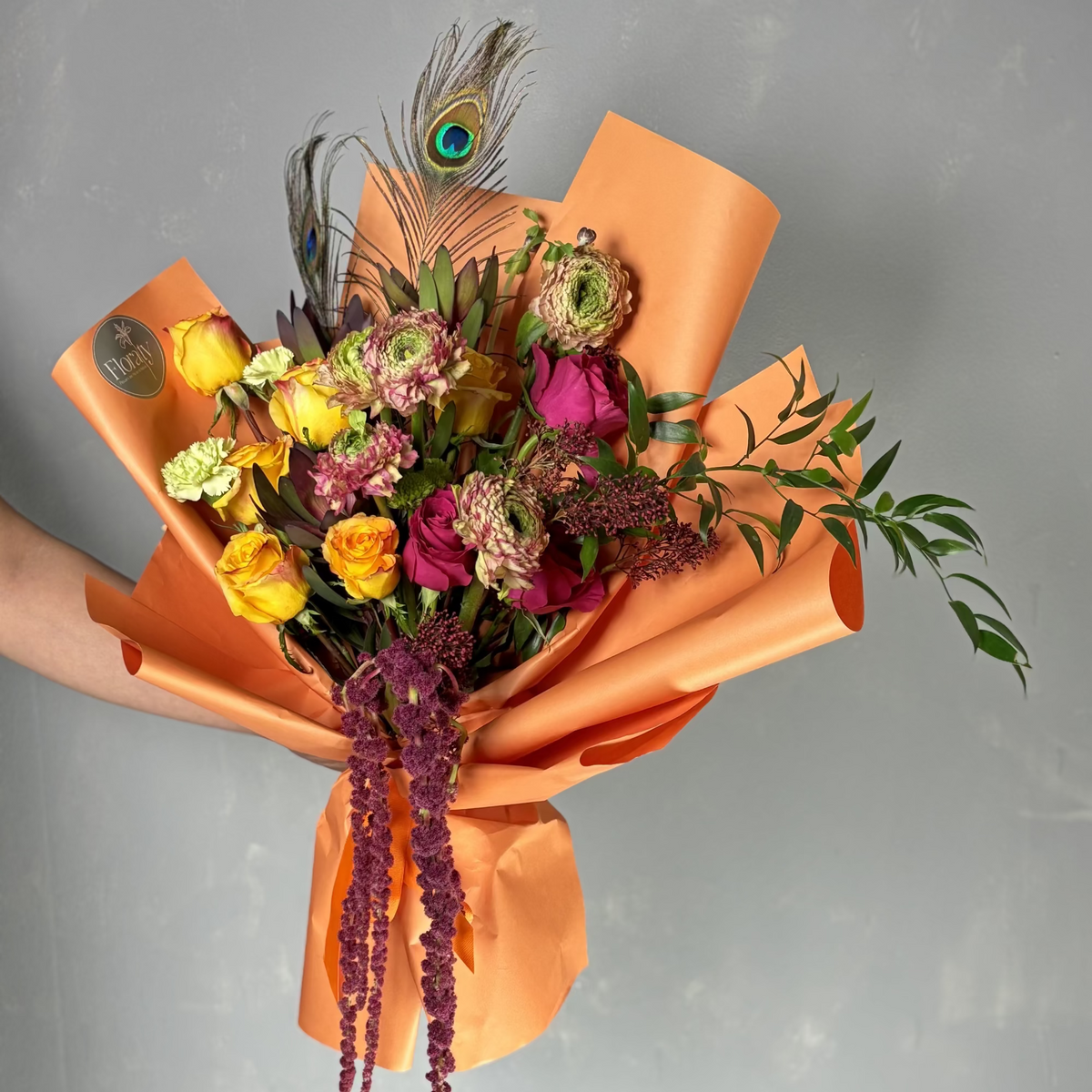 Bouquet of flowers wrapped in orange paper with a peacock feather on a gray background

