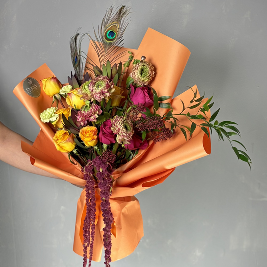 Bouquet of flowers wrapped in orange paper with a peacock feather on a gray background

