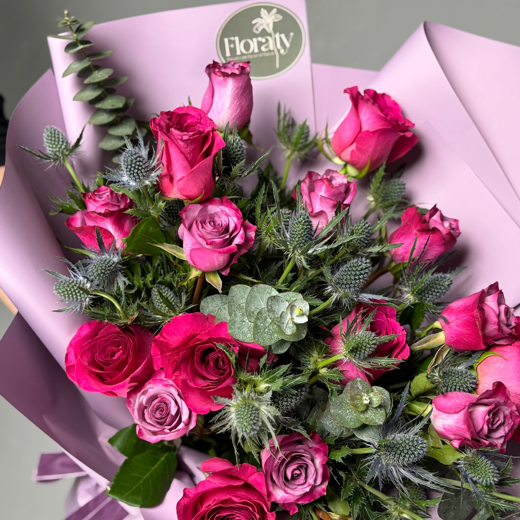 Bouquet of pink roses with greenery, wrapped in purple paper, on a light gray background.