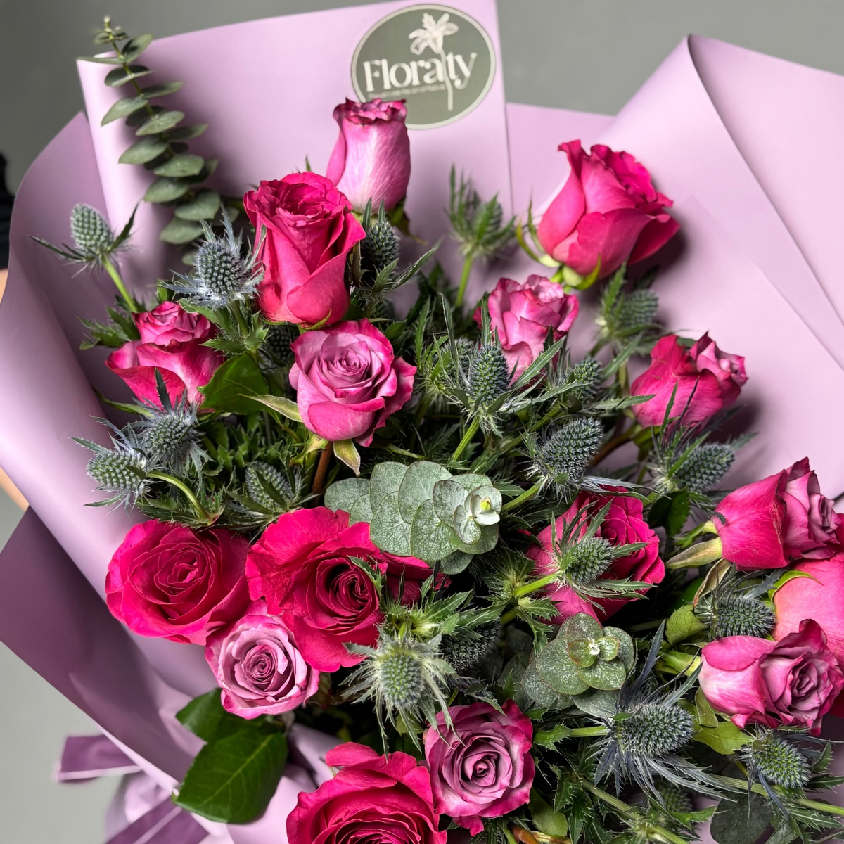 Bouquet of pink roses with greenery, wrapped in purple paper, on a light gray background.