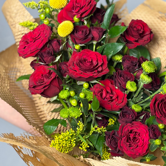 Bouquet of red roses with greenery and yellow flowers wrapped in brown paper.