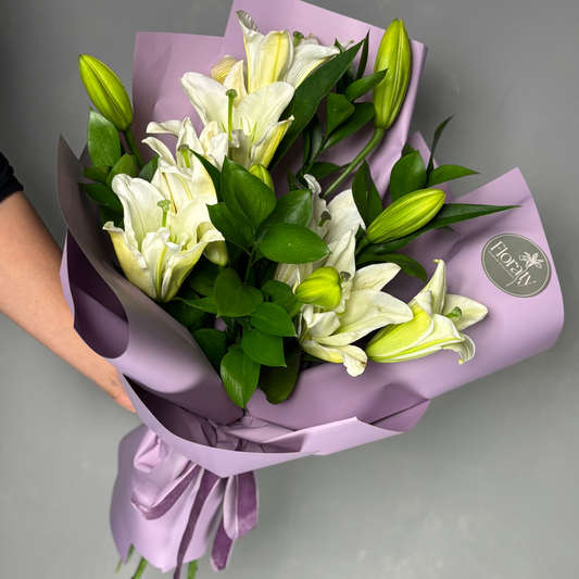 Bouquet of white lilies wrapped in purple paper with a visible brand logo on a gray background