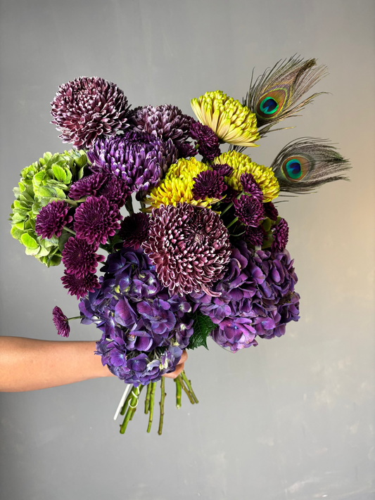 Bouquet of purple and yellow flowers with peacock feathers held by a person against a gray background