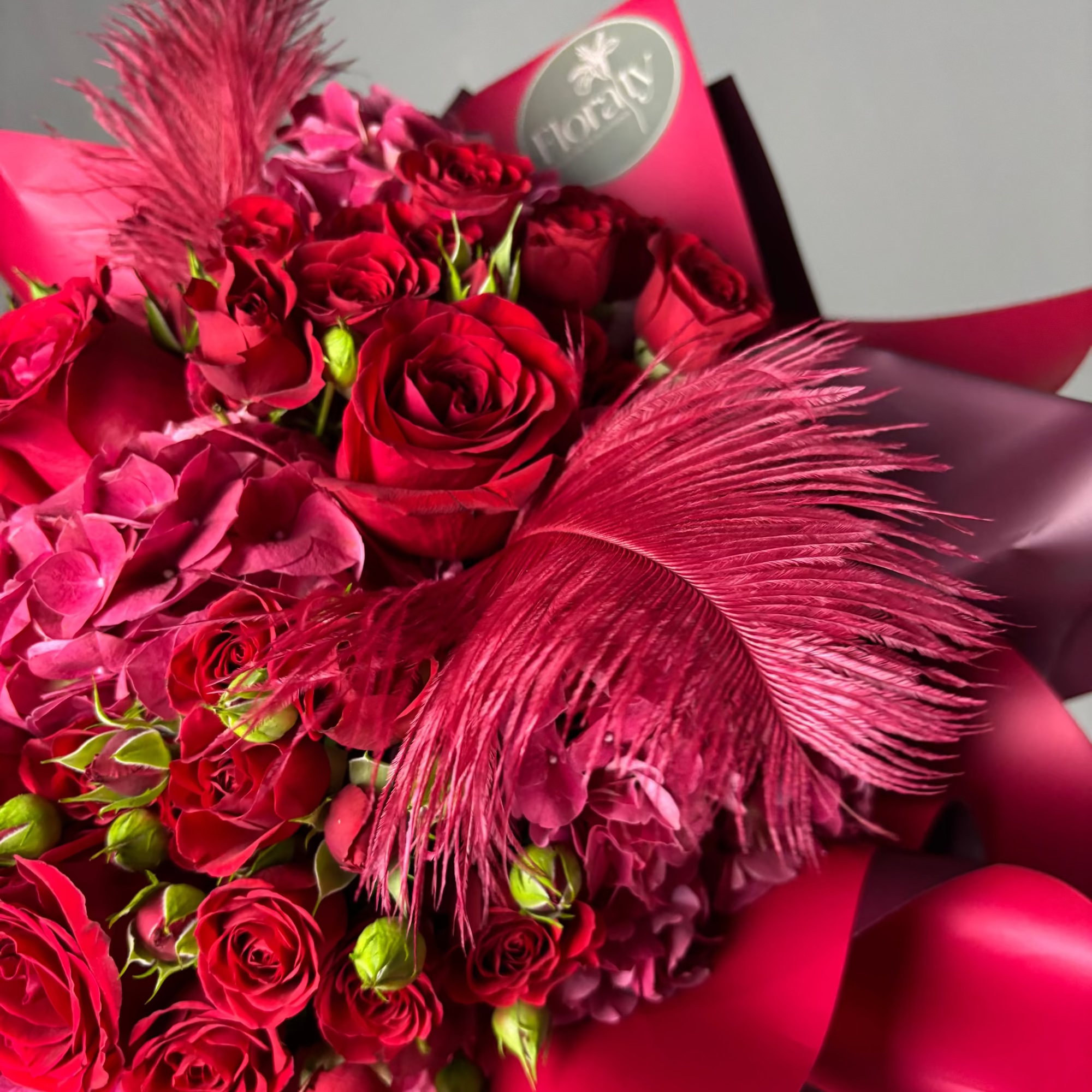 Bouquet of red roses with pink feathers and a visible brand logo on a gray background