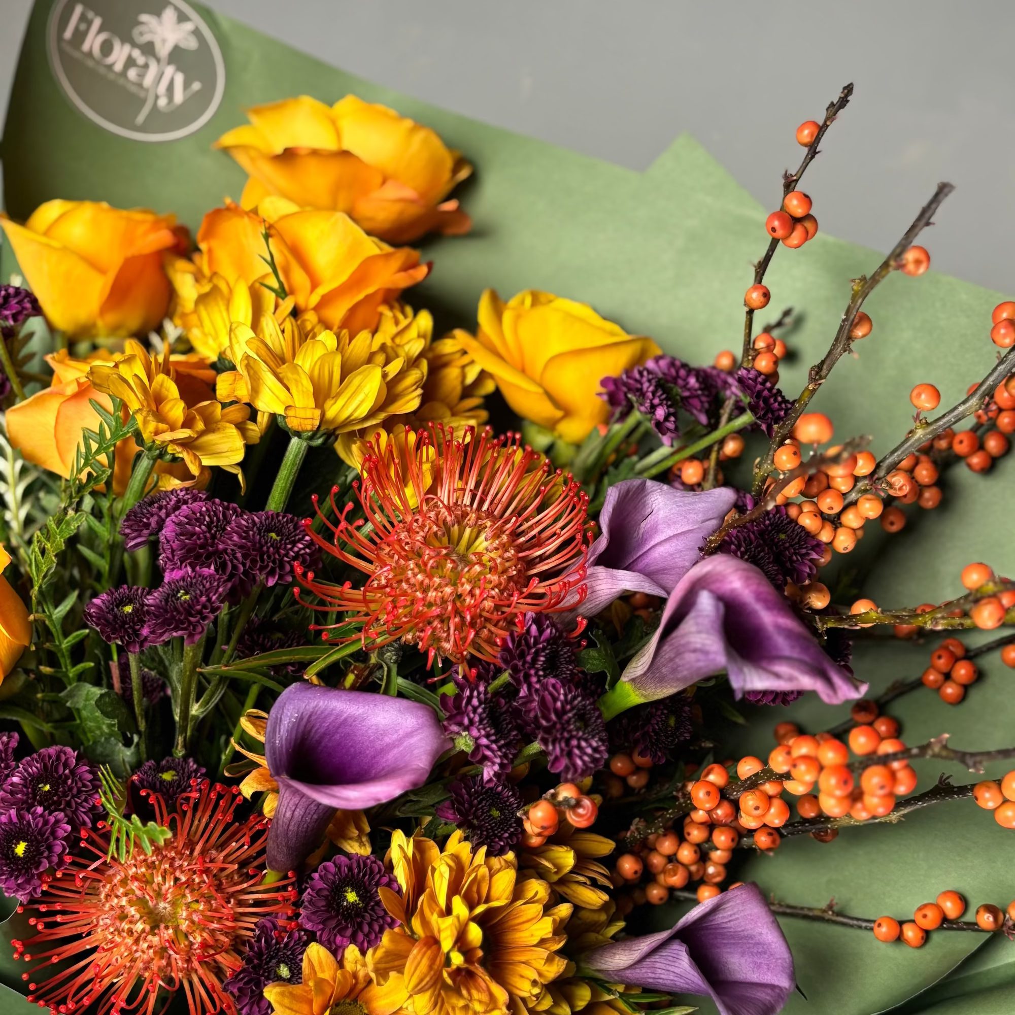 Bouquet of flowers with yellow, orange, and purple flowers on green paper, featuring the Floraty logo.